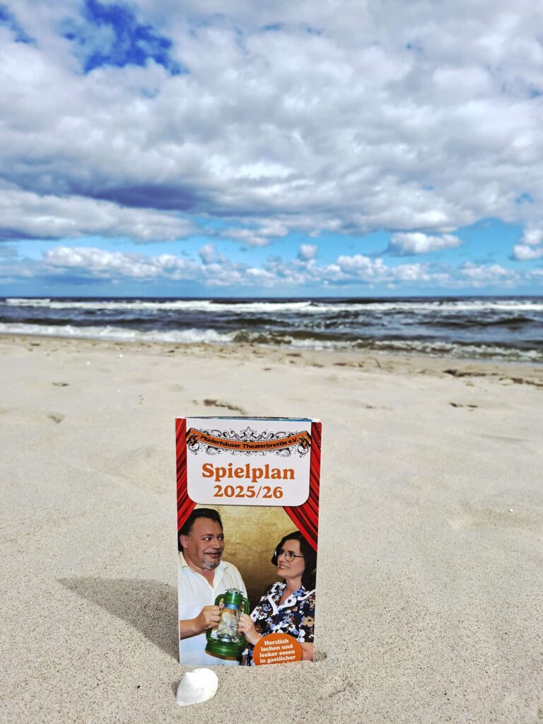 Das Bild zeigt ein kleines, aufgestelltes Heft mit der Aufschrift „Spielplan 2025/26“ im Sand am Strand. Im Hintergrund sind das Meer und ein bewölkter, blauer Himmel zu sehen.

Auf dem Cover des Heftes sind zwei Personen abgebildet, ein Mann und eine Frau. Der Mann hält einen großen Bierkrug in der Hand. Die Frau trägt eine Brille und ein buntes, gemustertes Oberteil. Der Titel des Heftes lautet „Spielplan 2025/26“, und darunter steht ein Text.

Der Sand ist im Vordergrund detailreich zu erkennen, und links neben dem Heft liegt eine kleine Muschel. Das Bild scheint an einem sonnigen, aber windigen Tag aufgenommen worden zu sein, was sich in den Wellen und der Wolkenformation widerspiegelt.
