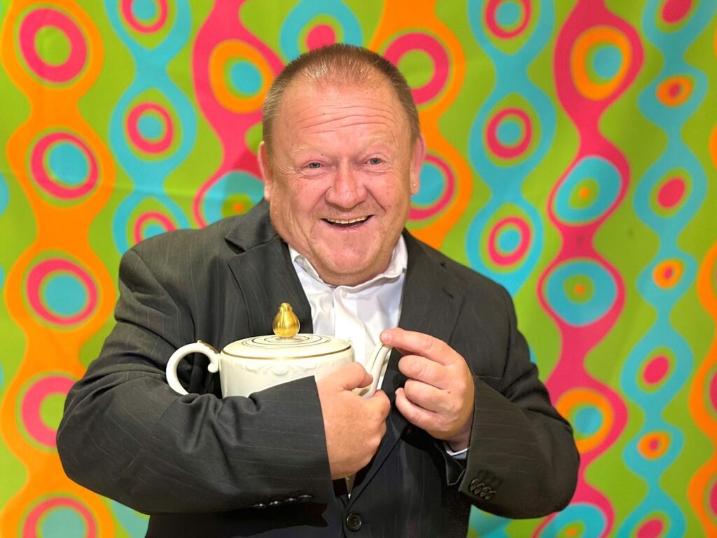 Das Bild zeigt einen lachenden Mann in einem dunklen Anzug mit weißem Hemd. Er hält eine weiße Teekanne mit goldenem Deckel in seinen Händen. Im Hintergrund ist eine bunte Tapete mit kreisförmigen, geschwungenen Mustern in leuchtenden Farben wie Grün, Türkis, Pink und Orange zu sehen. Die Beleuchtung scheint von vorne zu kommen, wodurch der Mann und die Teekanne gut ausgeleuchtet sind.