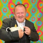 Das Bild zeigt einen lachenden Mann in einem dunklen Anzug mit weißem Hemd. Er hält eine weiße Teekanne mit goldenem Deckel in seinen Händen. Im Hintergrund ist eine bunte Tapete mit kreisförmigen, geschwungenen Mustern in leuchtenden Farben wie Grün, Türkis, Pink und Orange zu sehen. Die Beleuchtung scheint von vorne zu kommen, wodurch der Mann und die Teekanne gut ausgeleuchtet sind.