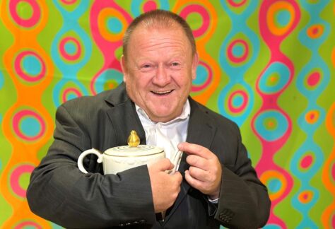 Das Bild zeigt einen lachenden Mann in einem dunklen Anzug mit weißem Hemd. Er hält eine weiße Teekanne mit goldenem Deckel in seinen Händen. Im Hintergrund ist eine bunte Tapete mit kreisförmigen, geschwungenen Mustern in leuchtenden Farben wie Grün, Türkis, Pink und Orange zu sehen. Die Beleuchtung scheint von vorne zu kommen, wodurch der Mann und die Teekanne gut ausgeleuchtet sind.
