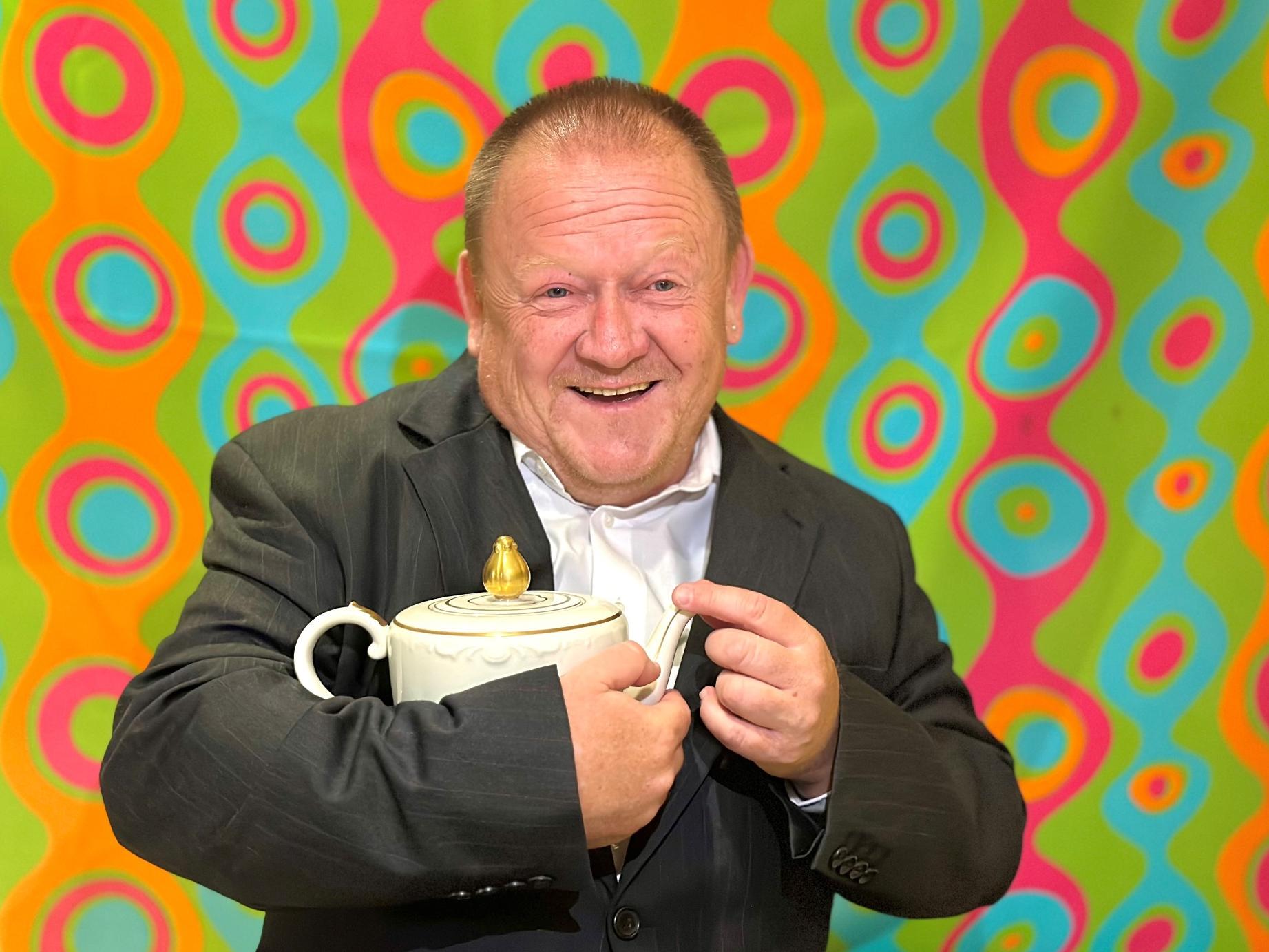 Das Bild zeigt einen lachenden Mann in einem dunklen Anzug mit weißem Hemd. Er hält eine weiße Teekanne mit goldenem Deckel in seinen Händen. Im Hintergrund ist eine bunte Tapete mit kreisförmigen, geschwungenen Mustern in leuchtenden Farben wie Grün, Türkis, Pink und Orange zu sehen. Die Beleuchtung scheint von vorne zu kommen, wodurch der Mann und die Teekanne gut ausgeleuchtet sind.