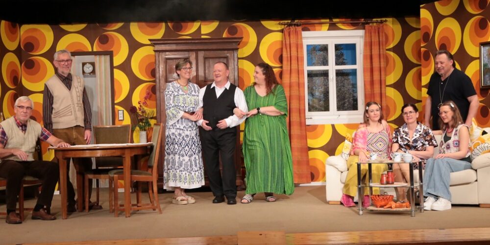 Brettle-Lampengeischd-alle 🎭 Barrierefreie Bildbeschreibung: Eine Szene aus einem Theaterstück Dies ist eine Aufnahme einer Theaterbühne, die ein Wohnzimmer im Stil der 1970er Jahre darstellt. Eine Gruppe von acht Erwachsenen, vier Männer und vier Frauen, steht oder sitzt in dieser Szene. 🖼️ Hintergrund und Setting Die Tapete ist sehr auffällig und dominiert den Hintergrund. Sie hat ein psychedelisches Muster aus großen, ineinander verschachtelten Ringen oder Kreisen in den Farben Braun, Gelb und Orange. In der Mitte der Bühne steht ein großer, dunkler, geschnitzter Kleiderschrank (möglicherweise ein Bauernschrank). Links vom Schrank führt eine Tür (oder ein Durchgang) in einen anderen Raum. Rechts vom Schrank befindet sich ein weißes Sprossenfenster, durch das eine nächtliche oder bewaldete Szene zu sehen ist. An den Wänden hängen zwei gerahmte, dunkle Gemälde (eines links, eines rechts), die Landschaften oder abstrakte Muster zeigen. 🛋️ Requisiten und Möbel Auf der linken Seite befindet sich ein dunkler, rechteckiger Esstisch mit zwei Stühlen. Auf der rechten Seite steht eine cremefarbene oder hellgelbe Couch mit passenden Kissen. Davor steht ein kleiner, glasbedeckter Tisch. Auf dem gläsernen Tisch stehen Tassen, Untertassen und eine kleine Schale mit rot-orangenen Gegenständen, möglicherweise Krebse oder Deko-Artikel. 👥 Die Personen Insgesamt sind acht Personen zu sehen, die in lockeren Gruppen angeordnet sind: Ganz links: Ein älterer Mann mit Brille und einem weißen Vollbart sitzt auf einem Stuhl links vom Esstisch. Er trägt ein helles, ärmelloses Oberteil (Weste) über einem karierten Hemd und dunkle Hosen. Hinter dem Tisch: Ein weiterer älterer Mann, steht lächelnd. Er trägt ein beiges Hemd oder Weste über einem hellen Oberteil und braune Hosen. Gruppe Mitte (stehend): Eine Frau steht links von der Mitte und lächelt. Sie trägt ein langes, knöchellanges Kleid mit einem blauen und weißen Muster. Ein Mann steht in der Mitte, mit den Armen um die beiden Frauen links und rechts von ihm. Er trägt eine schwarze Hose und eine schwarze Weste über einem weißen Hemd. Eine Frau steht rechts von ihm und lächelt. Sie trägt ein langes, locker fallendes grünes Kleid mit weiten Ärmeln. Gruppe Rechts (sitzend und stehend): Drei Frauen sitzen eng auf der Couch. Die Frau ganz links auf der Couch trägt ein pinkes/rosafarbenes Top mit Blumenmuster. Die mittlere Frau trägt ein dunkles, gemustertes Kleid. Die Frau ganz rechts trägt ein helles, langärmeliges Top und hellblaue Jeans. Ein Mann steht rechts, hinter der Couch. Er trägt ein dunkles (wahrscheinlich schwarzes) Poloshirt und eine dunkle Hose. Er lächelt und hat die Hand auf die Schulter der ganz rechten Frau auf der Couch gelegt. Die Beleuchtung ist hell, was auf eine Innenaufnahme hindeutet, wobei der Hintergrund stark durch die Tapete und die Dunkelheit durch das Fenster kontrastiert wird. Die Kleidung der Darsteller variiert zwischen traditionell und zeitgenössisch, was typisch für eine Komödie oder ein Volkstheaterstück sein könnte.