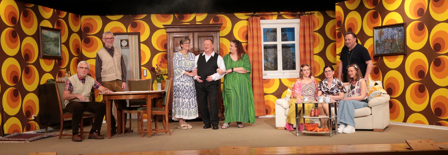 🎭 Barrierefreie Bildbeschreibung: Eine Szene aus einem TheaterstückDies ist eine Aufnahme einer Theaterbühne, die ein Wohnzimmer im Stil der 1970er Jahre darstellt. Eine Gruppe von acht Erwachsenen, vier Männer und vier Frauen, steht oder sitzt in dieser Szene.

🖼️ Hintergrund und Setting

    Die Tapete ist sehr auffällig und dominiert den Hintergrund. Sie hat ein psychedelisches Muster aus großen, ineinander verschachtelten Ringen oder Kreisen in den Farben Braun, Gelb und Orange.

    In der Mitte der Bühne steht ein großer, dunkler, geschnitzter Kleiderschrank (möglicherweise ein Bauernschrank).

    Links vom Schrank führt eine Tür (oder ein Durchgang) in einen anderen Raum.

    Rechts vom Schrank befindet sich ein weißes Sprossenfenster, durch das eine nächtliche oder bewaldete Szene zu sehen ist.

    An den Wänden hängen zwei gerahmte, dunkle Gemälde (eines links, eines rechts), die Landschaften oder abstrakte Muster zeigen.

🛋️ Requisiten und Möbel

    Auf der linken Seite befindet sich ein dunkler, rechteckiger Esstisch mit zwei Stühlen.

    Auf der rechten Seite steht eine cremefarbene oder hellgelbe Couch mit passenden Kissen. Davor steht ein kleiner, glasbedeckter Tisch.

    Auf dem gläsernen Tisch stehen Tassen, Untertassen und eine kleine Schale mit rot-orangenen Gegenständen, möglicherweise Krebse oder Deko-Artikel.

👥 Die Personen

Insgesamt sind acht Personen zu sehen, die in lockeren Gruppen angeordnet sind:

    Ganz links: Ein älterer Mann mit Brille und einem weißen Vollbart sitzt auf einem Stuhl links vom Esstisch. Er trägt ein helles, ärmelloses Oberteil (Weste) über einem karierten Hemd und dunkle Hosen.

    Hinter dem Tisch: Ein weiterer älterer Mann, steht lächelnd. Er trägt ein beiges Hemd oder Weste über einem hellen Oberteil und braune Hosen.

    Gruppe Mitte (stehend):

        Eine Frau steht links von der Mitte und lächelt. Sie trägt ein langes, knöchellanges Kleid mit einem blauen und weißen Muster.

        Ein Mann steht in der Mitte, mit den Armen um die beiden Frauen links und rechts von ihm. Er trägt eine schwarze Hose und eine schwarze Weste über einem weißen Hemd.

        Eine Frau steht rechts von ihm und lächelt. Sie trägt ein langes, locker fallendes grünes Kleid mit weiten Ärmeln.

    Gruppe Rechts (sitzend und stehend):

        Drei Frauen sitzen eng auf der Couch.

            Die Frau ganz links auf der Couch trägt ein pinkes/rosafarbenes Top mit Blumenmuster.

            Die mittlere Frau trägt ein dunkles, gemustertes Kleid.

            Die Frau ganz rechts trägt ein helles, langärmeliges Top und hellblaue Jeans.

        Ein Mann steht rechts, hinter der Couch. Er trägt ein dunkles (wahrscheinlich schwarzes) Poloshirt und eine dunkle Hose. Er lächelt und hat die Hand auf die Schulter der ganz rechten Frau auf der Couch gelegt.

Die Beleuchtung ist hell, was auf eine Innenaufnahme hindeutet, wobei der Hintergrund stark durch die Tapete und die Dunkelheit durch das Fenster kontrastiert wird. Die Kleidung der Darsteller variiert zwischen traditionell und zeitgenössisch, was typisch für eine Komödie oder ein Volkstheaterstück sein könnte.