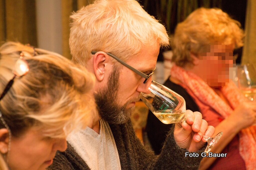 „Nahaufnahme einer Weinverkostung in warmer Atmosphäre. Ein Mann mit blondem Haar, Bart und Brille hält ein Glas Weißwein an die Nase und riecht konzentriert an den Aromen. Er ist im Profil von links zu sehen. Im Hintergrund und Anschnitt sind weitere Teilnehmer der Probe erkennbar. Das Licht ist golden und gemütlich.“